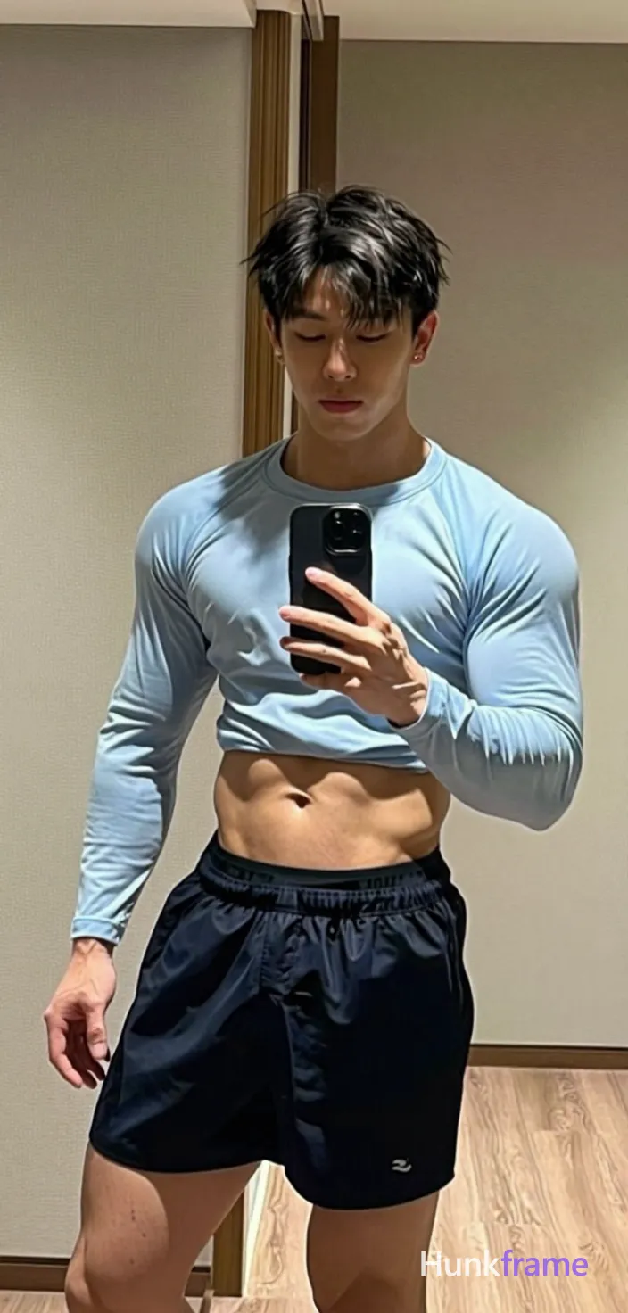 Asian man with light to medium tan skin and short black textured hair, clean-shaven face holding a vertical black smartphone against his chest in front of a full-length mirror; He wears a light blue long-sleeve compression shirt over dark navy athletic shorts revealing black briefs at the waistband; Athletic build with defined abs stands upright on light wood flooring with beige walls and a wooden door frame behind him, slight leftward lean captured by camera angle; Soft ambient indoor lighting reflects evenly on skin; composition includes slight fisheye distortion typical of phone cameras, shot on iPhone 15 Pro main camera, authentic smartphone computational photography, natural Smart HDR and true-to-life skin texture, vertical 9:16 Xiaohongshu lifestyle feed, bright clean everyday scene, unretouched phone photo with subtle sensor grain, trendy Chinese internet 网红 aesthetic, candid influencer energy, phone held naturally in one hand for selfie, phone visible in frame; single clean uninterrupted photograph with a cohesive editorial composition and plain continuous surrounding surfaces