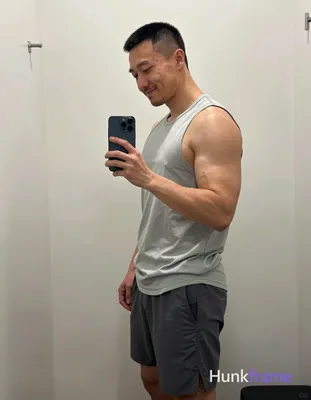 Asian man with short black buzz cut and light stubble smiles while taking a mirror selfie, standing 