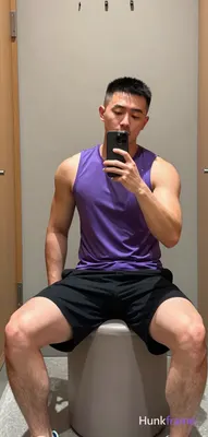 Asian man with short black buzz cut and light stubble sits on a cylindrical ottoman in a gym locker 